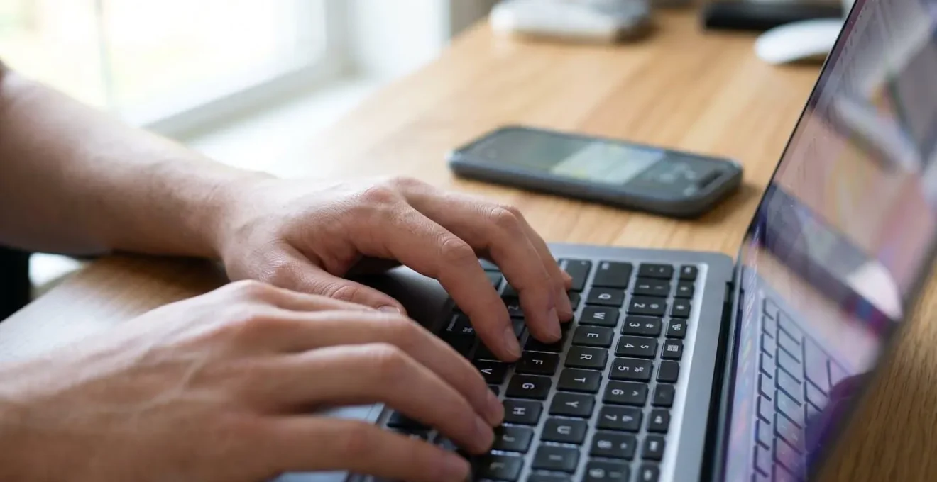 Close-up of hands typing on a laptop keyboard with a partially visible spreadsheet on screen and a smartphone showing notifications beside it